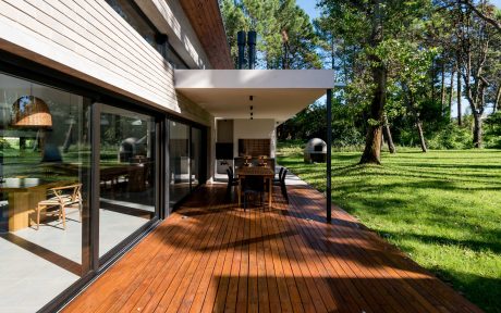 A modern wooden house with a large deck overlooking a well-manicured lawn and trees.
