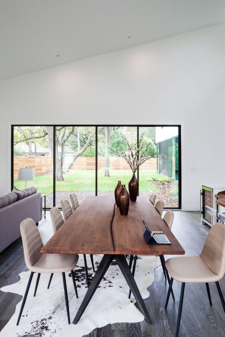 Minimalist dining room with large windows overlooking a lush, green backyard and distinctive wooden table.
