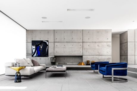 Modern living room with concrete walls, gray sofa, blue velvet chairs, and geometric coffee table.