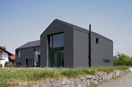 A modern, sleek two-story home with large windows, a black facade, and a stone wall.