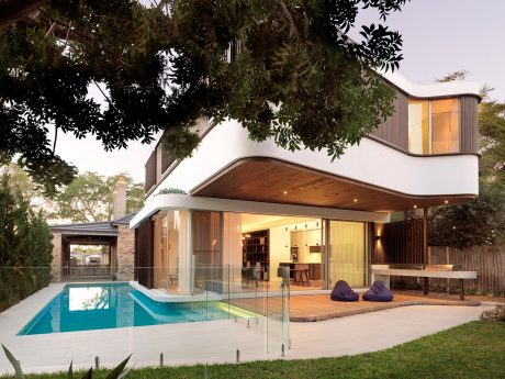 Modern, single-story house with glass walls, pool, and lush greenery surrounding the property.