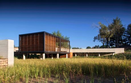 This modern structure features a wooden lattice facade and is elevated on concrete pillars, blending with the surrounding natural landscape.