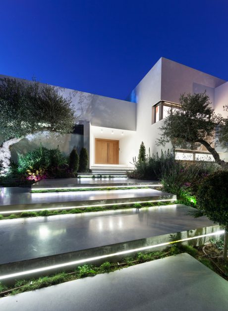 A modern, minimalist home with a striking facade, surrounded by lush landscaping and illuminated walkway.