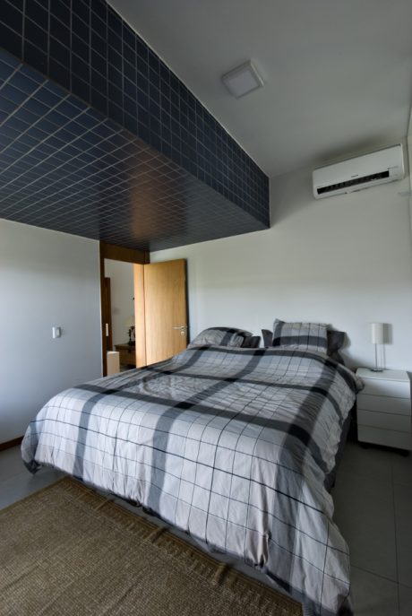 A modern bedroom with black tiled walls, white ceiling, and checkered bedding. An air conditioning unit is visible.