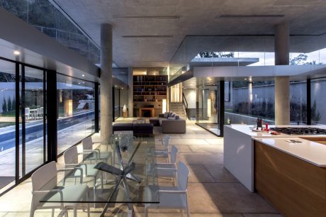 Spacious, contemporary open-plan living space with sleek glass walls, concrete beams, and minimalist decor.