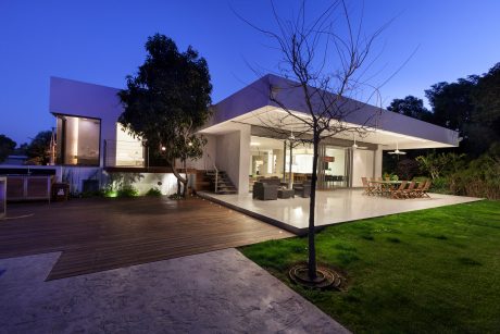 Sleek, modern home with a large wooden deck, glass walls, and landscaped yard at night.