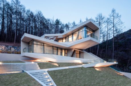 Modern, asymmetric villa with glass walls, surrounded by trees and a lawn with stairs.