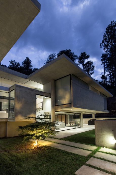 A modern, angular concrete building with glass walls and a sloped roof, surrounded by lush greenery.