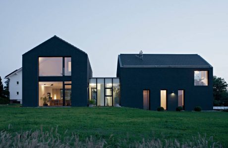 Modern black houses with large windows, surrounded by a grassy field at dusk.