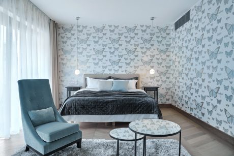 Modern bedroom with butterfly-patterned wallpaper, plush gray bedding, and teal accent chair.