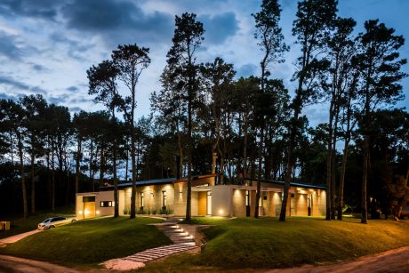 A modern, single-story building with large windows, illuminated at night and surrounded by a lush forest.