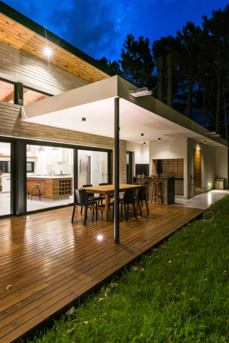 Modern, open-concept home with wooden deck, outdoor seating, and glass walls overlooking lush foliage.