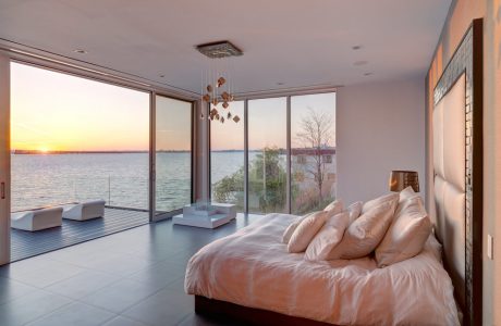 Spacious modern bedroom with floor-to-ceiling windows overlooking a lake at sunset.