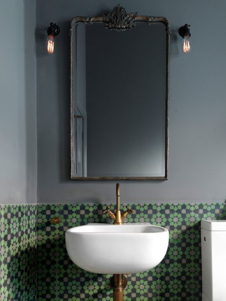 Ornate antique mirror with wall sconces over a tile-covered vanity with brass fixtures.