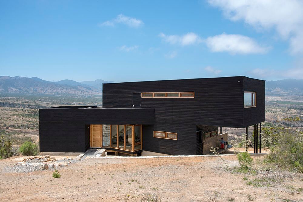 A modern black wooden structure with large windows overlooking a mountainous landscape.