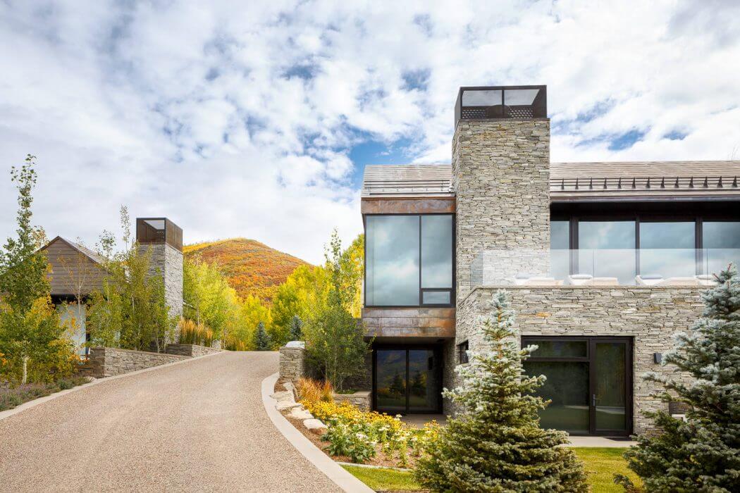 A contemporary stone and glass residence nestled among autumn-hued trees, with a paved driveway leading to the entrance.