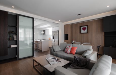 Minimalist living room with modern kitchen visible through glass partition, warm wooden accents.