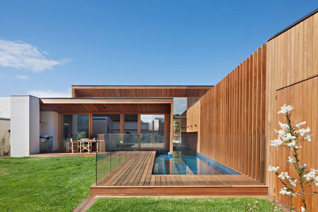 Modern wooden exterior with covered patio, glass walls, and a rectangular pool on a lawn.