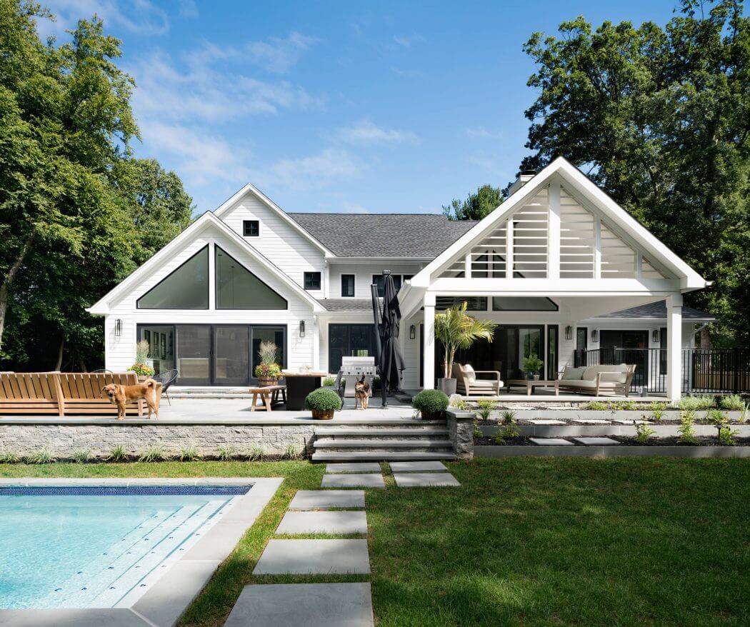 Large white house with gabled roof, wrap-around porch, and modern outdoor entertaining area.