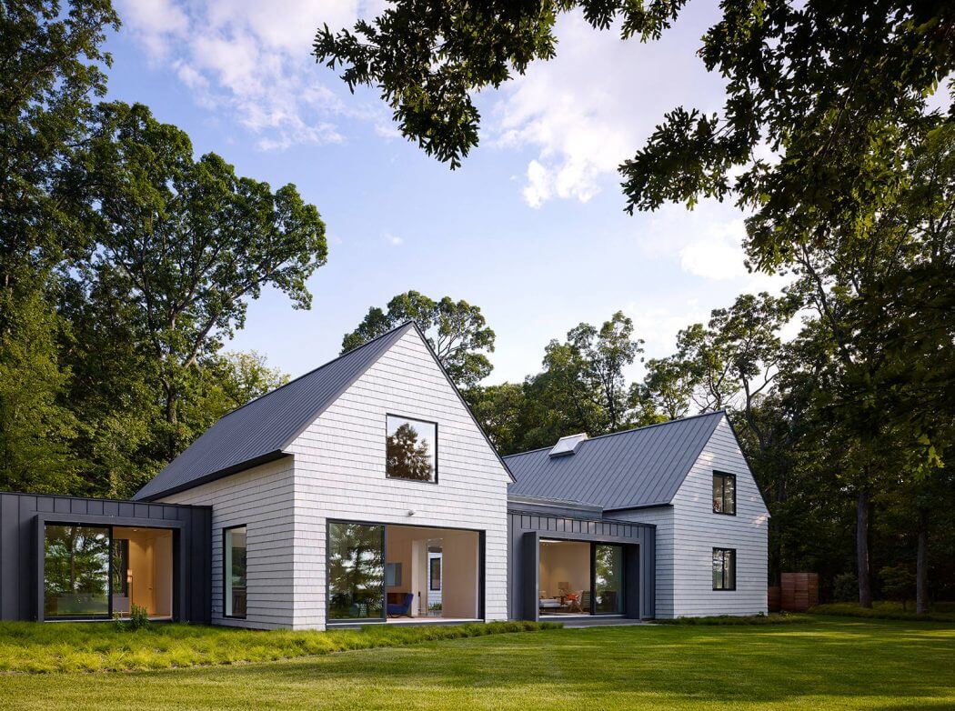 Modern farmhouse-style home with gabled roofs, large windows, and a lush green lawn surrounded by mature trees.