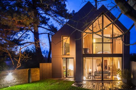 Cozy modern cabin with large windows, wooden deck, and lush foliage at night.