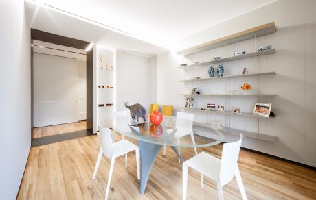 Sleek, airy dining area with modern glass table, white chairs, and open shelving displaying decor.