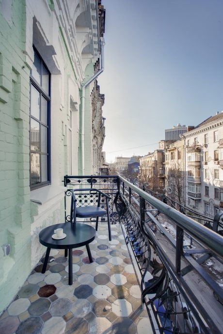 A cozy balcony with a wrought-iron railing, stone tile floor, and cafe-style furnishings against a backdrop of historic buildings.