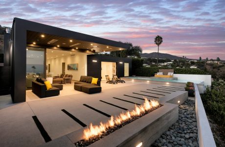 An elegant modern outdoor living space with a sleek, illuminated fire feature, surrounding seating, and a distant palm tree under a colorful sunset sky.