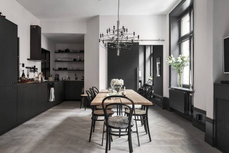 A modern, minimalist kitchen and dining area featuring a large wooden table, black chairs, and a striking chandelier.