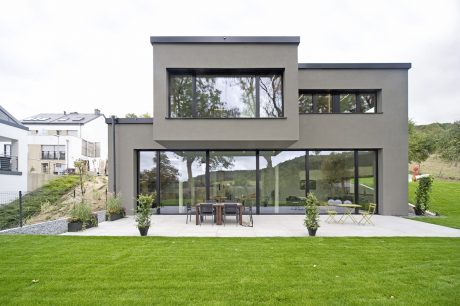 Contemporary residential home with large glass windows, patio furniture, and lush green lawn.