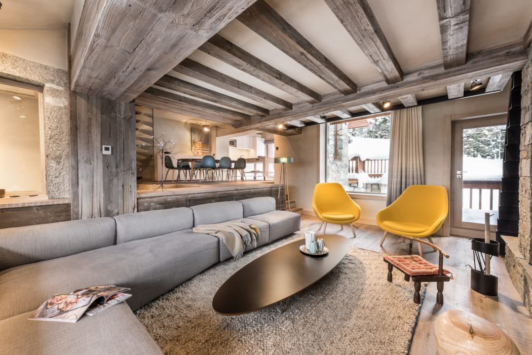 Rustic chalet with exposed wooden beams, gray sofa, yellow chairs, and oval coffee table.