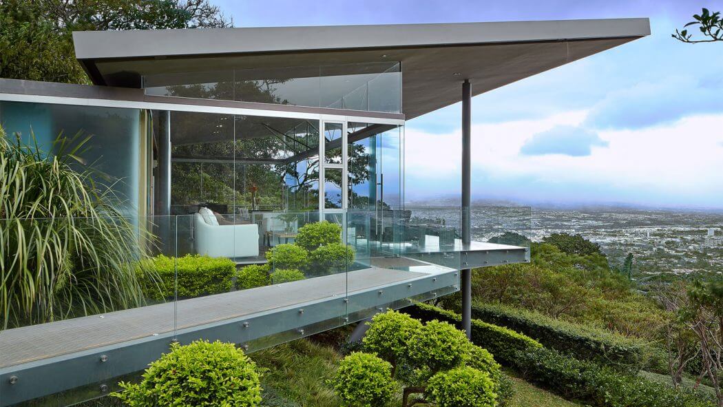 Modern glass pavilion with open floor plan, surrounded by lush greenery and expansive city view.