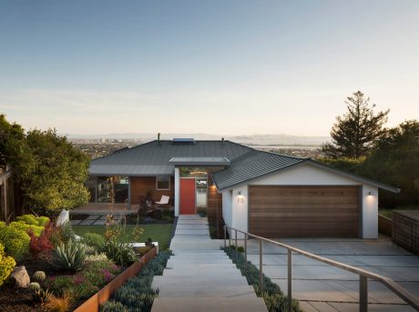 A modern home with a wooden exterior, a red door, and a rooftop surrounded by landscaping with a scenic view.