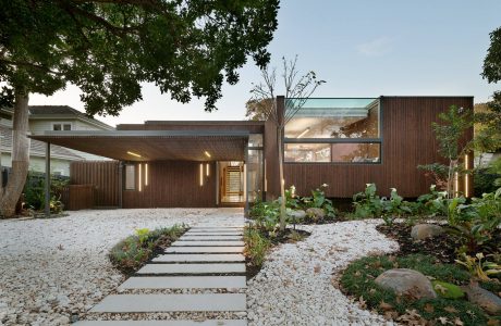A modern, wooden-clad residence with a covered entryway and lush landscaping.