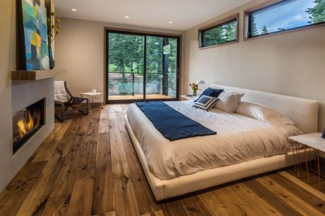 Spacious, modern bedroom with large windows showcasing lush forest scenery and fireplace.