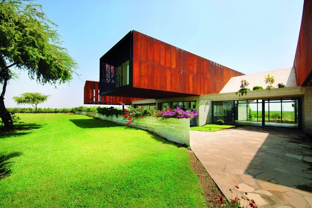 A modern, red-toned concrete building with a lush green lawn and colorful flowers in the foreground.