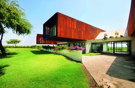 A modern, red-toned concrete building with a lush green lawn and colorful flowers in the foreground.