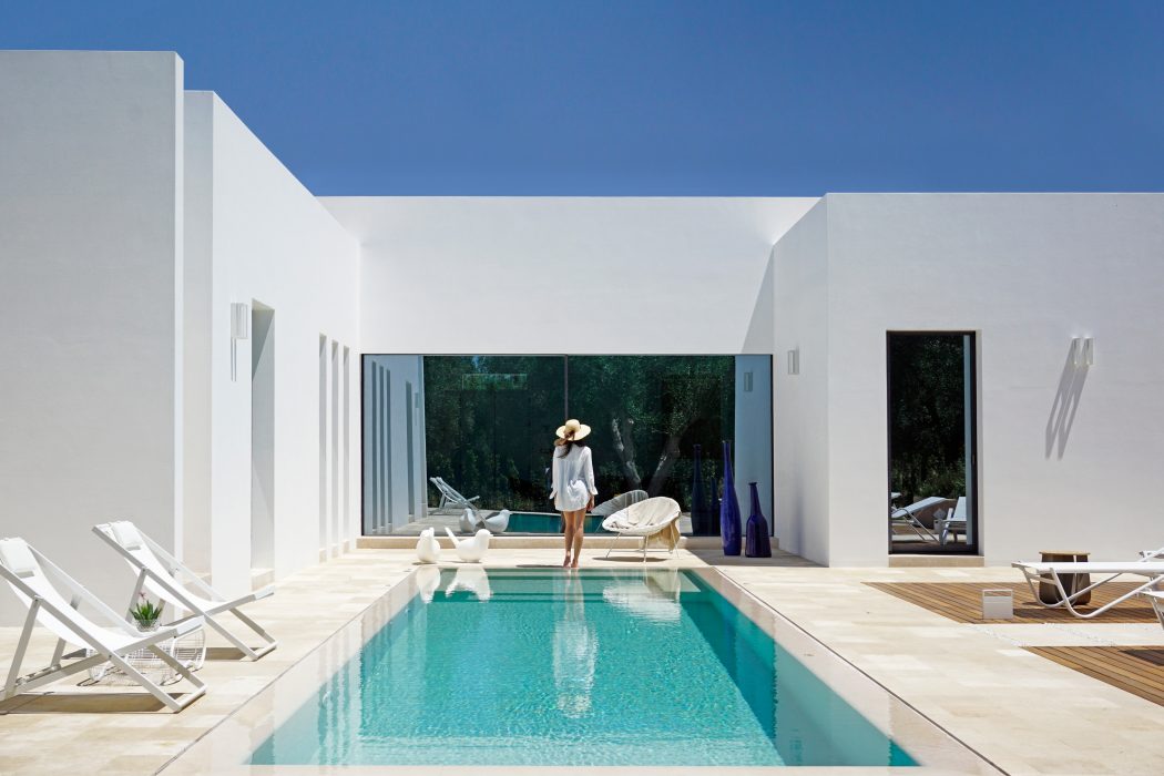 A modern, minimalist pool area with white walls, loungers, and a vibrant turquoise pool under a clear sky.