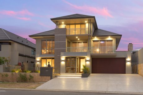 A modern, multi-story house with a prominent entryway, balconies, and large windows illuminated at night.