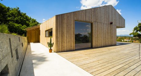 Wooden modern building with large windows and a spacious wooden deck overlooking greenery.
