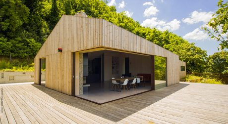 A modern, open-plan wooden structure with glass walls, surrounded by lush greenery.