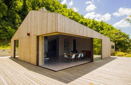 A modern, open-plan wooden structure with glass walls, surrounded by lush greenery.