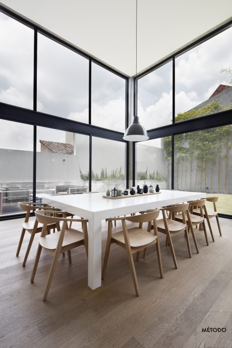 A contemporary open-plan dining area with a large white table, wooden chairs, and tall windows offering scenic views.