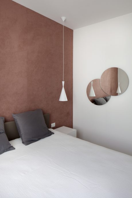A minimalist bedroom with a warm accent wall, pendant lights, and a round mirror.
