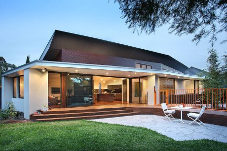 Modern open-concept home with wooden deck, large windows, and lush landscaping.