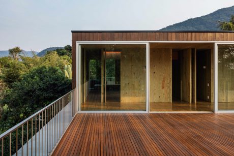 A modern wooden cabin with large glass windows and a spacious balcony overlooking lush greenery.