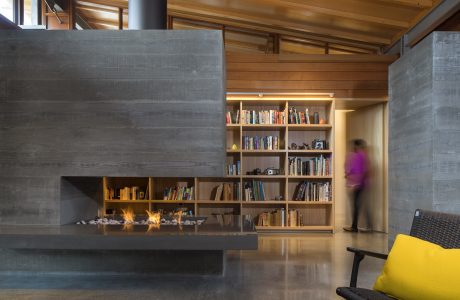 Warm, modern interior features a large concrete fireplace, bookshelves, and a blurred figure.