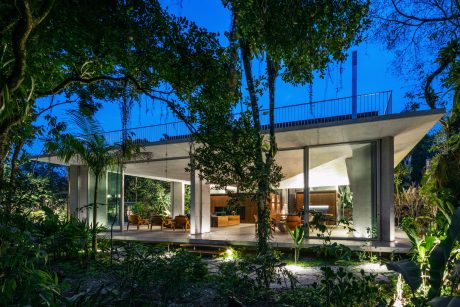 A modern, glass-walled house with an expansive roof and lush, surrounding vegetation.