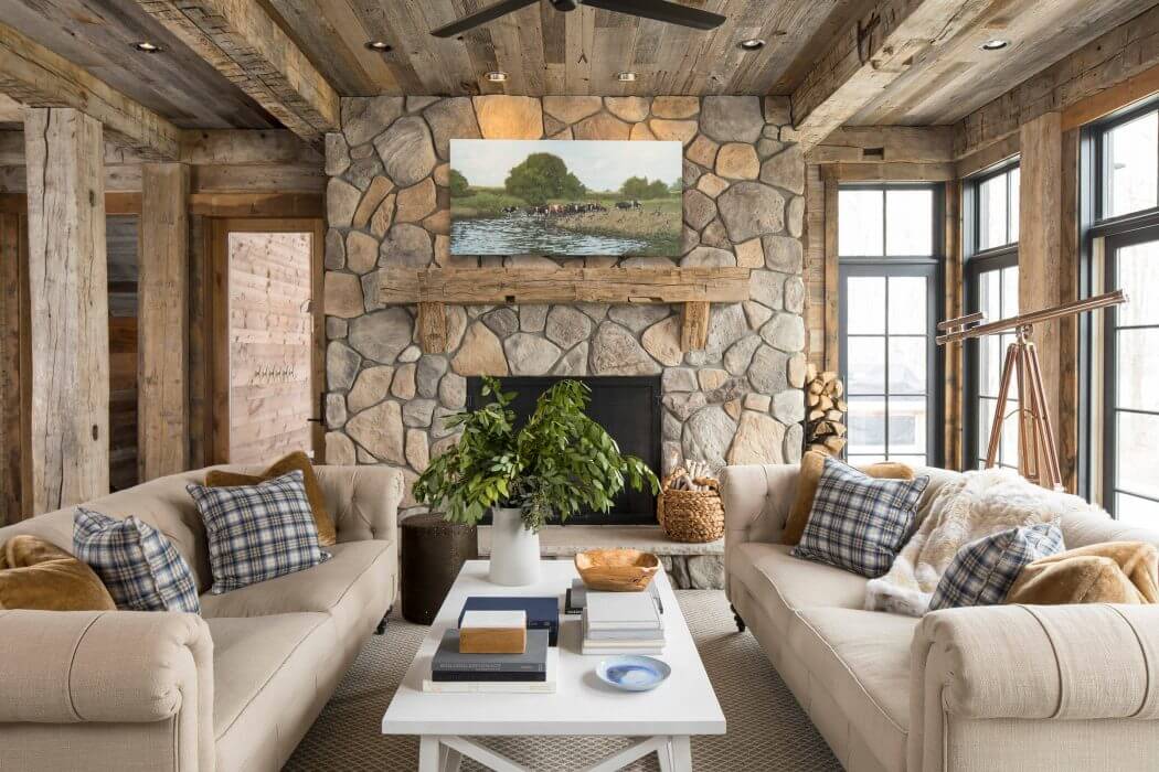 Rustic cabin interior with stone fireplace, wooden beams, and plaid-covered sofas.