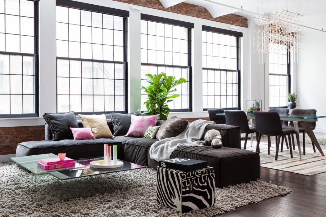 Spacious loft-style living room with large windows, black sofas, and modern chandelier.